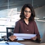 Focused young businesswoman reading financial report document and using a laptop in the workplace.