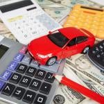 Red toy car surrounded by four calculators and a red pencil sitting on top of U.S. currency to represent the fees required when purchasing a car.