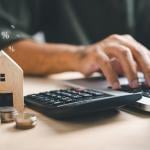 A person using a laptop and calculator with a wooden figure of a house and coins beside him.