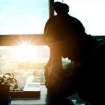 Silhouete of person sitting at desk in front of laptop, with head in hands backlit by sunset shining through office window in background.