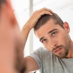 Young man with shaved head looking at mirror in bathroom at home, running fingers over hair.. 