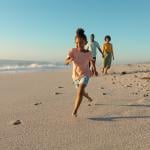 A young girl running on sand against parents walking on a beach.