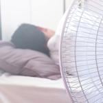 A white electric fan in the foreground in front of a person sleeping in bed, facing away.