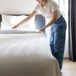 Low angle view of fresh white sheets tucked into bed with person backlit by window fixing the head of the bed.