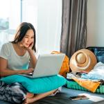 Young woman using a laptop beside an unpacked suitcase preparing for a trip.