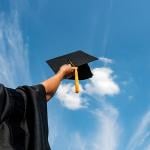 A graduate student holding up a graduation cap in the sky.