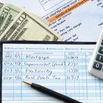 Balancing a checkbook with U.S. bills and calculator and bills visible on a table.