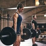 Strong woman and in foreground lifting bar bell with weights with man in background gym.