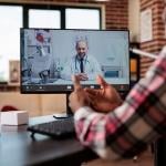 Computer screen showing telehealth call with male balding doctor sitting in office with stethoscope around his neck, while person dressed in plaid long sleeve shirt in foreground converses on video call with his back to the camera.