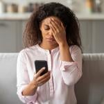 A photo capturing the tension of a young African American woman sitting on her couch, visibly overwhelmed as she looks at her smartphone in a neatly arranged living room.