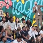 Side view of rows of high school students sitting on bleachers listening to a speaker wearing a tan suit talking into a mic.