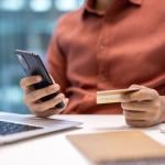 Businessperson holding a credit card while using a phone and laptop at an office desk.