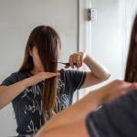 A woman cuts her hair in front of a mirror to have bangs.