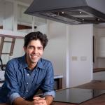 Adrian Grenier stands beneath his island-mounted CopperSmith® range hood, featuring a smooth bronze finish, paired with a handcrafted wood countertop for a warm, natural aesthetic.