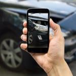 Person photographing passenger side headlight of damaged black car with smart phone for accident insurance.