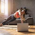Woman doing a side plank pose on a mat on a wooden floor, following a video on a laptop, with sunlight coming in through large window in the background. 