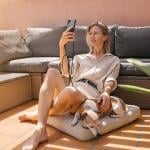 Woman wearing neutral button up top and shorts sitting on a floor cushion leaning against a couch, next to her dog, facetiming on her phone.