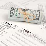 A tied up roll of hundred dollar bills on top of various paper U.S. tax forms like the 1040 and 1041.