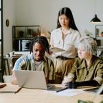 Three diverse young adults gathered around a laptop in a modern office setting.
