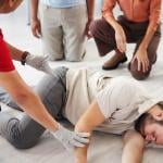 Demonstration by person teaching class for first aid, with someone as victim lying on their side and three people kneeling and watching.