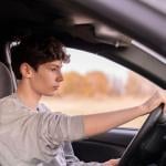 Young male driver holding steering wheel in car seen through the passenger side window.