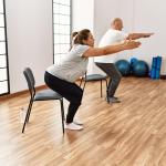 Two people each squatting toward a chair with their arms out in front of them, in a yoga studio type room.