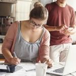 Woman doing calculations at kitchen table with laptop open and pen and paper while man with face not visible in the background is on his phone.