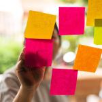 7 Pink, orange and yellow post-it notes stuck on glass with person whose face is obscured, writing on them from the other side.