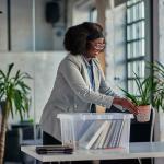 Black woman with glasses placing plants on an office desk with a clear plastic bin full of belongings on table.