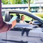 Arm of person in focus in the foreground, holding smartphone, with blurred dark car pulling up in the background, driver visible through the passenger window.