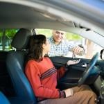 A teenage daughter inside a car receiving its keys from her proud dad.
