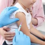 closeup of child's arm with vaccine needle held next to it by a person with blue gloves, an adult in a pink shirt holds child from other side.