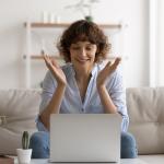 A happy woman sitting on her couch reading good news on her laptop.