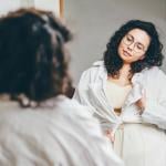 A curvy woman stands in front of a mirror, styling her outfit with a long sleeve shirt.