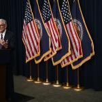 Fed chair Jerome Powell speaking at a podium with six flags behind him, 3 U.S. flags and 3 Board of Governors flags in alternating pattern.