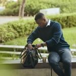 A young black businessman, looking inside his bag to eat lunch while sitting on a bench at a park.