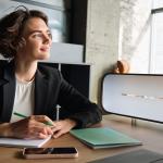 Business woman in office looks toward window thinking while taking notes.