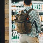 A man traveling with a backpack facing the cold drinks section of an airport food store.