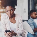 A black couple on their individual phones while sitting apart on the same couch at home.