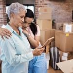 An older woman looking at a photograph while getting a hug from a younger family member while packing boxes on moving day.