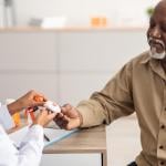 Portrait of black medical worker holding personal alarm button and giving it to mature patient, sitting at table in office, explaining how to use medical alert.
