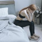 A young woman sits by her bed while pensively checking weight on a scale.