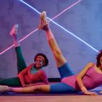 Two young adults wearing 80s style fitness clothing lay on their sides doing side leg raise exercise on floor at gym.