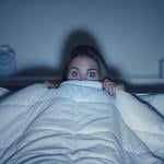 Stylized photo of woman hiding under covers with wide eyes, illuminated by screen from TV.