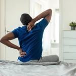 A young black man stretches and reaches for his back after getting up from bed.