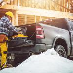 A construction worker in yellow gear prepares tools from a truck at a snowy building site with wooden building framing in the background.