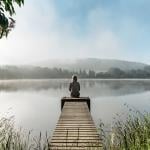 A woman sits quietly at the end of a small pier by a lake during a calm, misty morning.