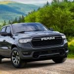 Black 2026 Ram 1500 REV Bighorn driving up dirt road with rolling green hills in background.