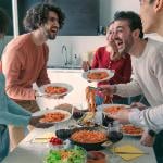 Five people gathered around a table standing with plates full of spaghetti.
