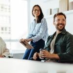 A group of three business people happily listens to a speaker during an office meeting.